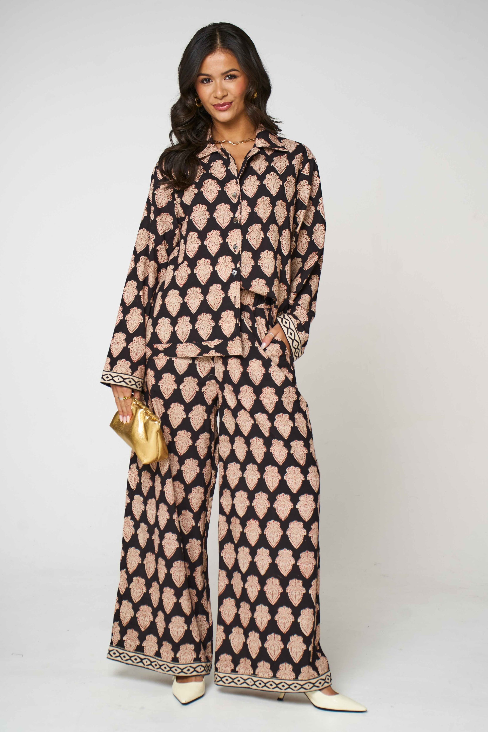 A woman modeling a matching two-piece coord set with a black and beige geometric pattern, featuring a long-sleeve button-up shirt and wide-leg pants, holding a small gold clutch against a light gray background.