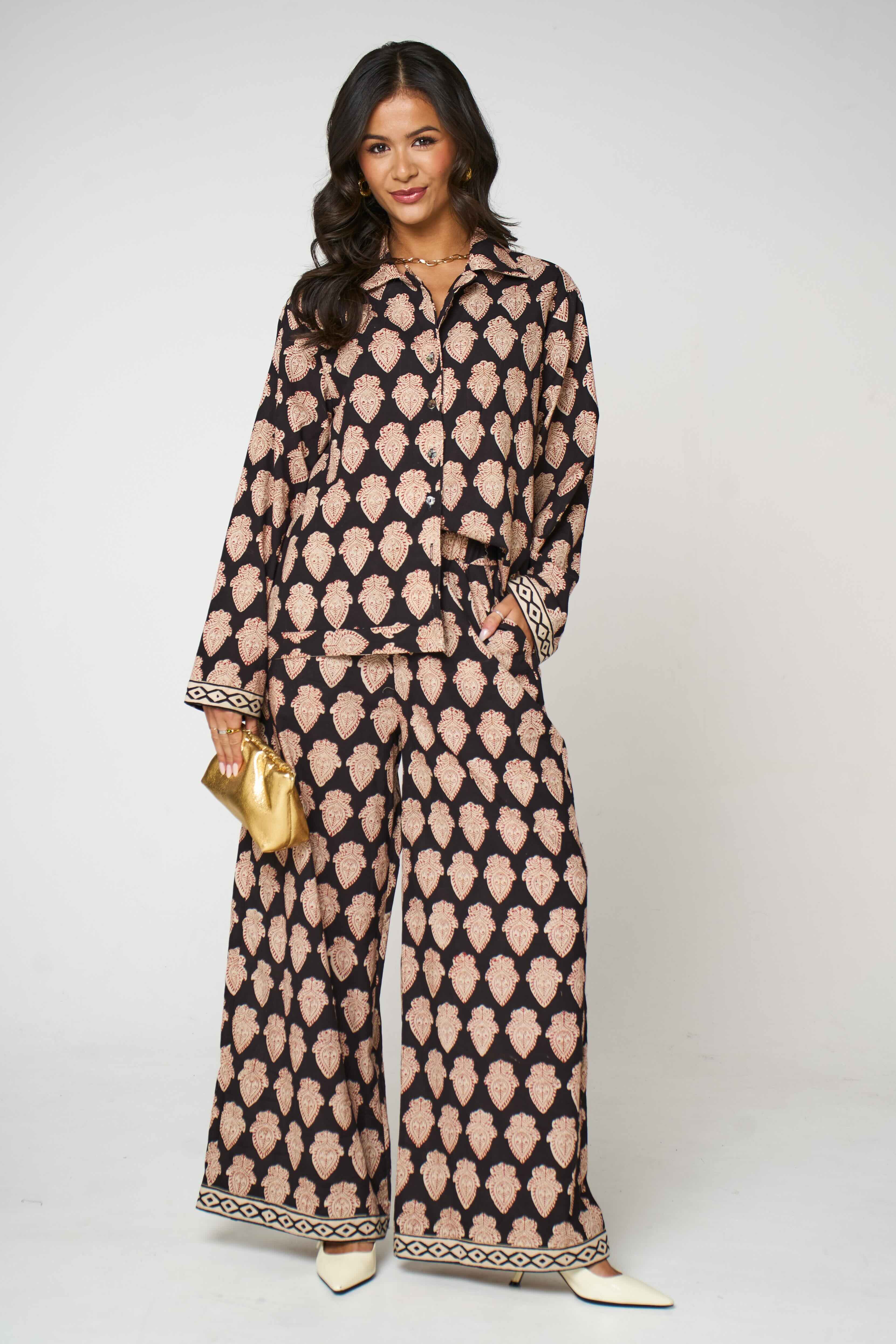 A woman modeling a matching two-piece coord set with a black and beige geometric pattern, featuring a long-sleeve button-up shirt and wide-leg pants, holding a small gold clutch against a light gray background.