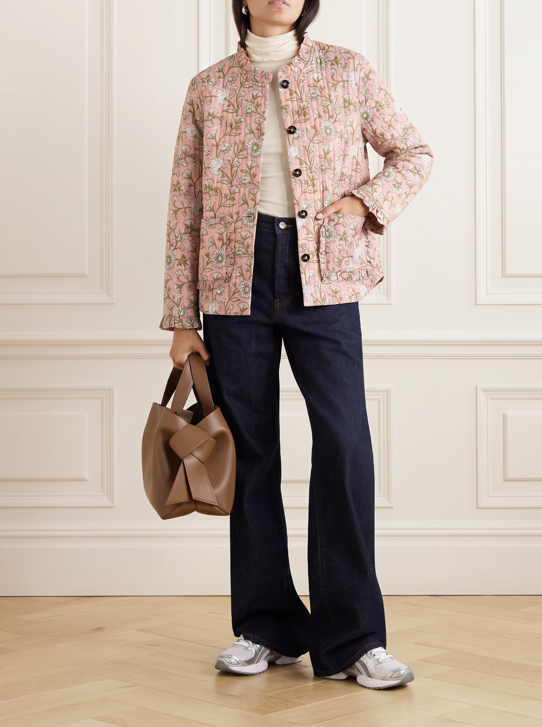 A model stands in a room with white panelled walls and a light wood floor, wearing a light pink block print quilted cotton floral jacket over a cream turtleneck, dark navy wide-leg jeans, and white sneakers, while holding a brown leather handbag.
