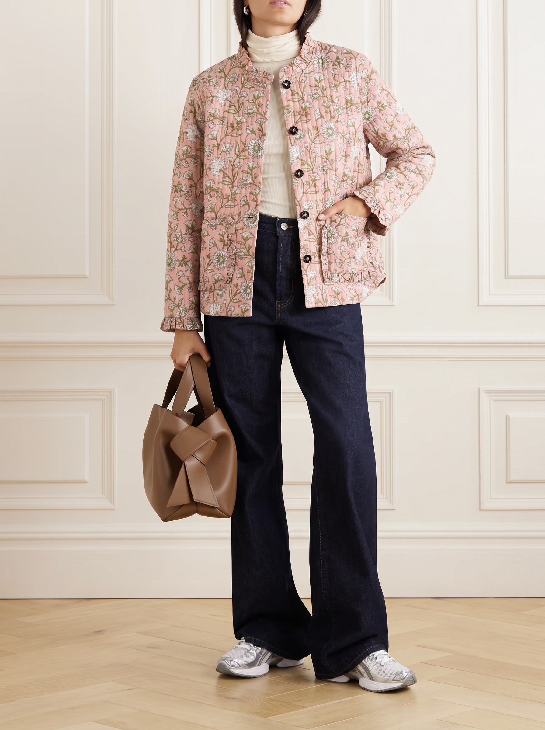 A model stands in a room with white panelled walls and a light wood floor, wearing a light pink block print quilted cotton floral jacket over a cream turtleneck, dark navy wide-leg jeans, and white sneakers, while holding a brown leather handbag.