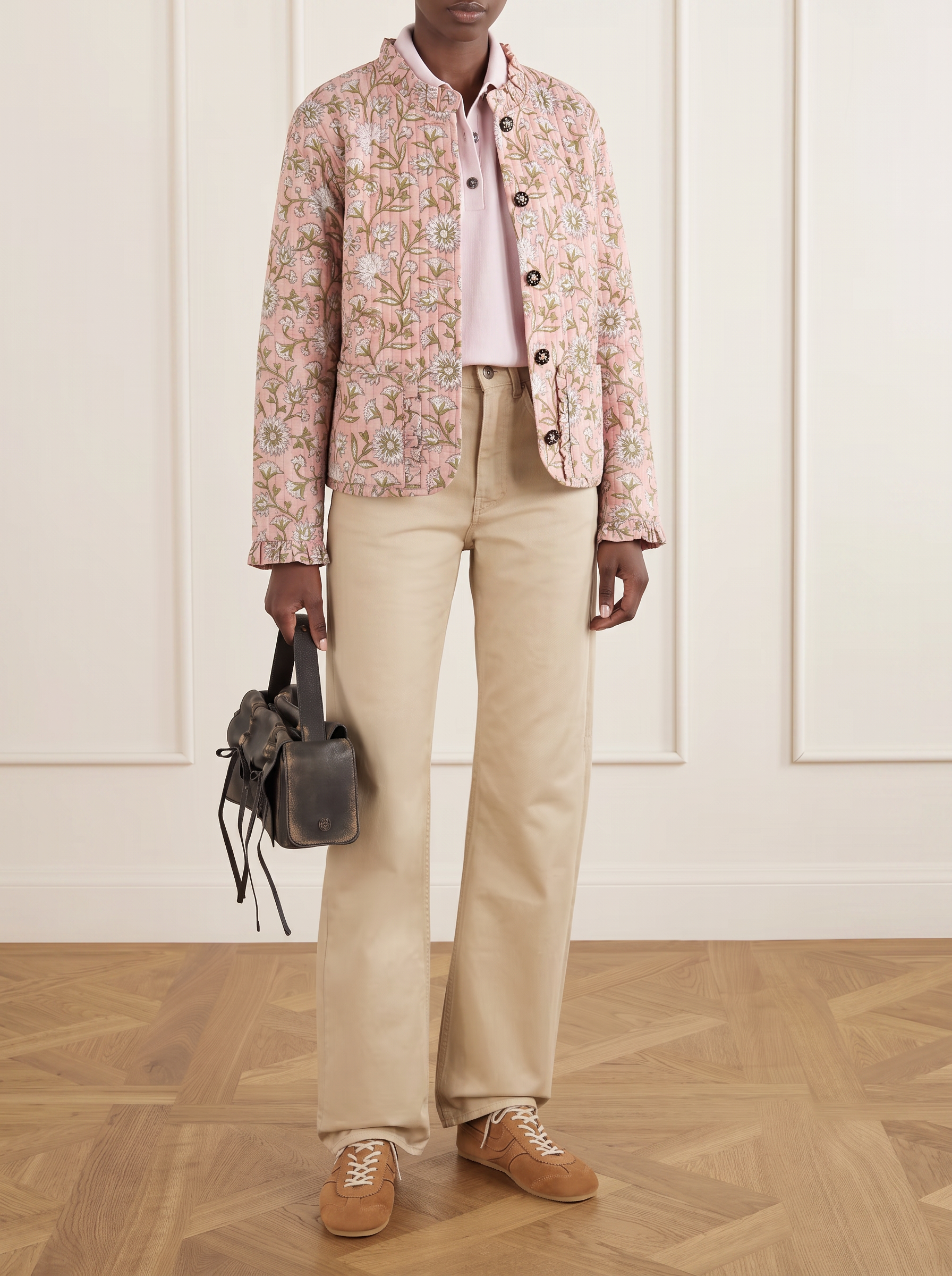 A model stands in a room with paneled cream walls and a wood parquet floor, wearing a reversible quilted light pink cotton block print floral patterned jacket over a pale pink polo shirt, beige wide-leg trousers, and tan suede shoes, while holding a dark gray leather handbag.