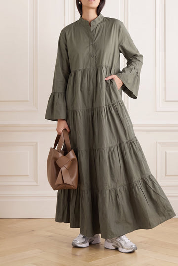A woman models a long-sleeved, tiered maxi dress in olive green, holding a brown leather handbag and wearing white sneakers, set against a classic cream paneled wall and light wood floor.