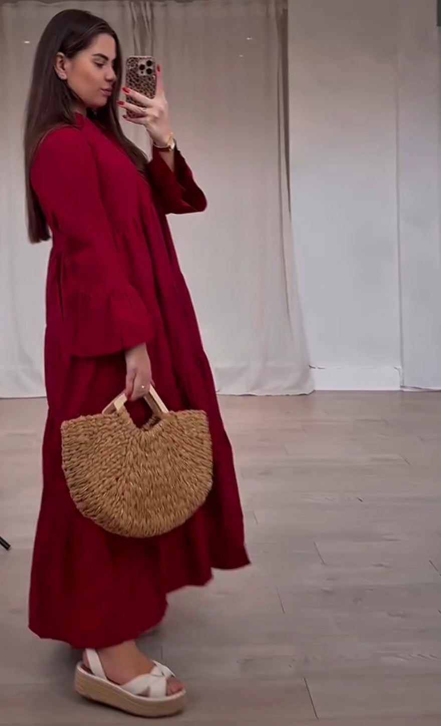 A woman takes a mirror selfie wearing a long-sleeved, loose-fitting maxi dress in deep red. She is holding a woven straw half-moon handbag with wooden handles and wearing white platform sandals.